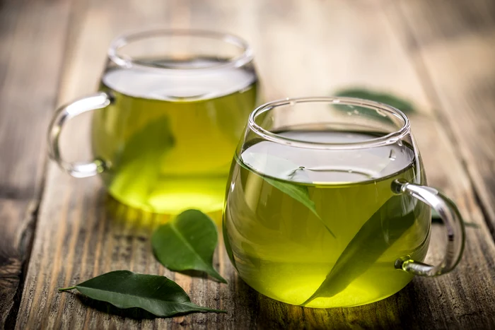 Two cups full of green tea and a few leaves of green tea in them and on the table on which they are 