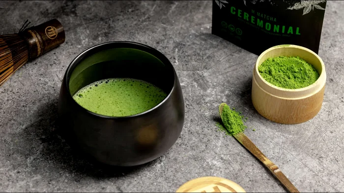 A box with green tea powder and a cup of green tea next to the utensils for youtube preparation