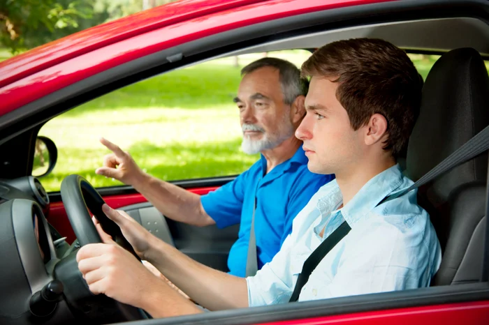 A young man at the wheel of a red car, along with an older instructor, with gray hair