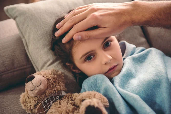 Little girl with fever, in bed with a teddy bear, parent with her hand on her forehead