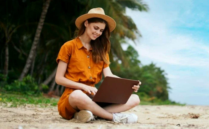 A young woman stands with the laptop on her knees, on a beach