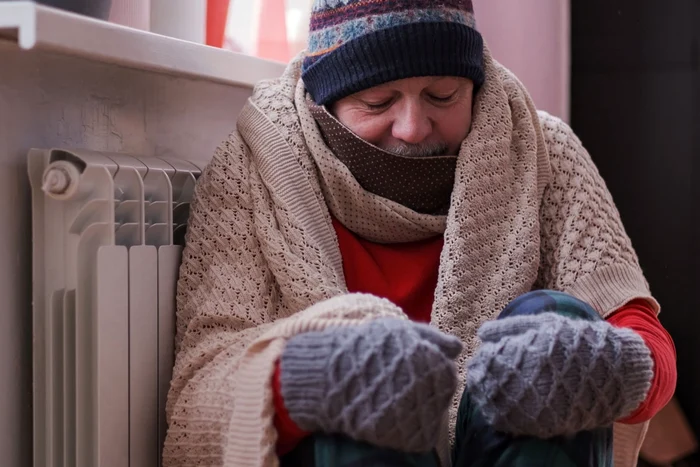 A very thickly dressed man is crouching next to the cold radiator